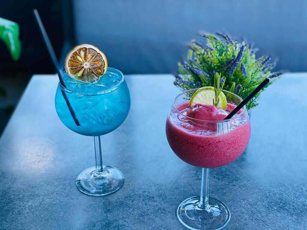 Două cocktailuri pe masă: unul albastru cu felie de lămâie uscată și unul roșu, frappé, cu lime