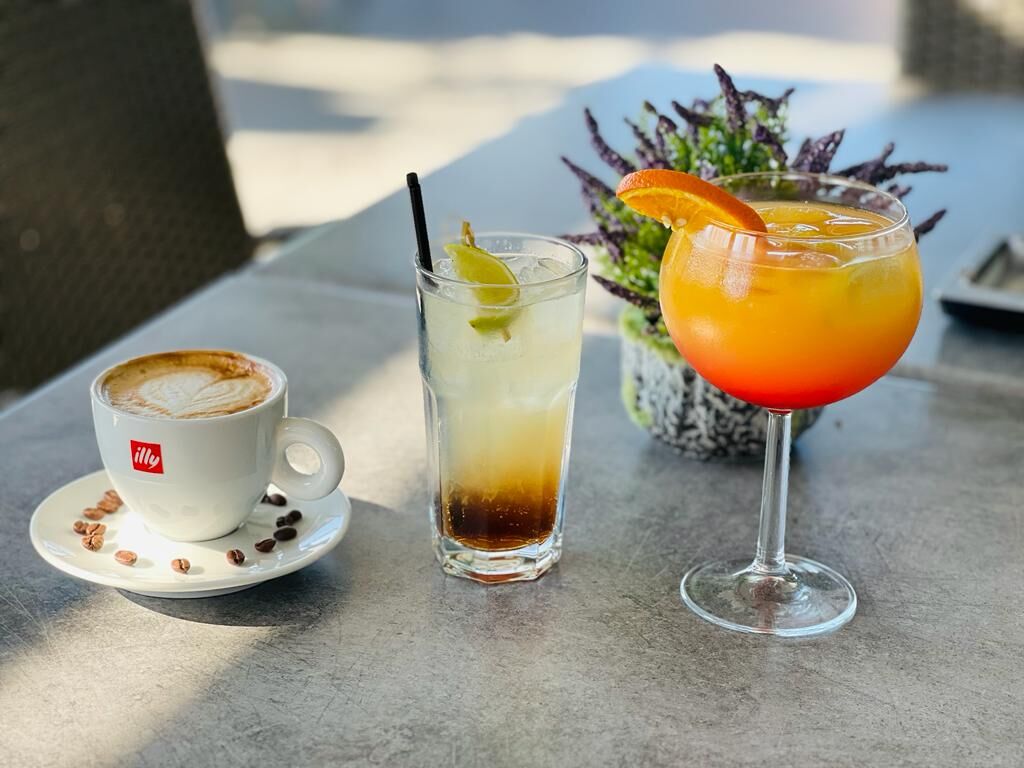 Cappuccino illy cu latte art, o băutură cu lămâie și un cocktail portocaliu pe masa de pe terasă